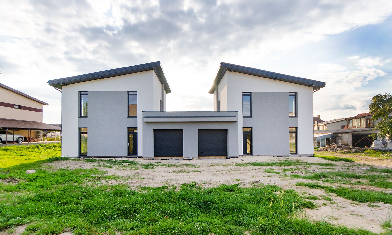 Newly built two-story side-by-side duplex with white and gray stucco walls, a shed roof on each unit and two dark garage doors in a shared center section, set on a bare dirt and grass lot under a cloudy sky