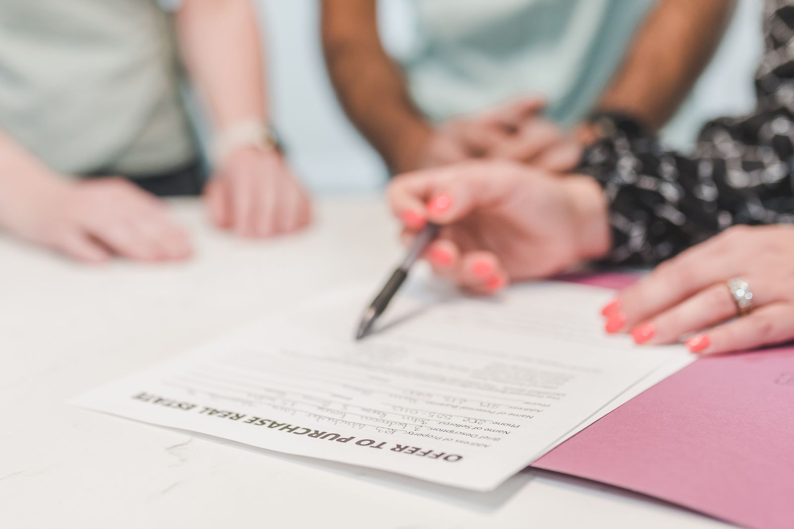 Three people seated at a white table over a printed offer-to-purchase real estate form, one of them holding a pen above the signature area