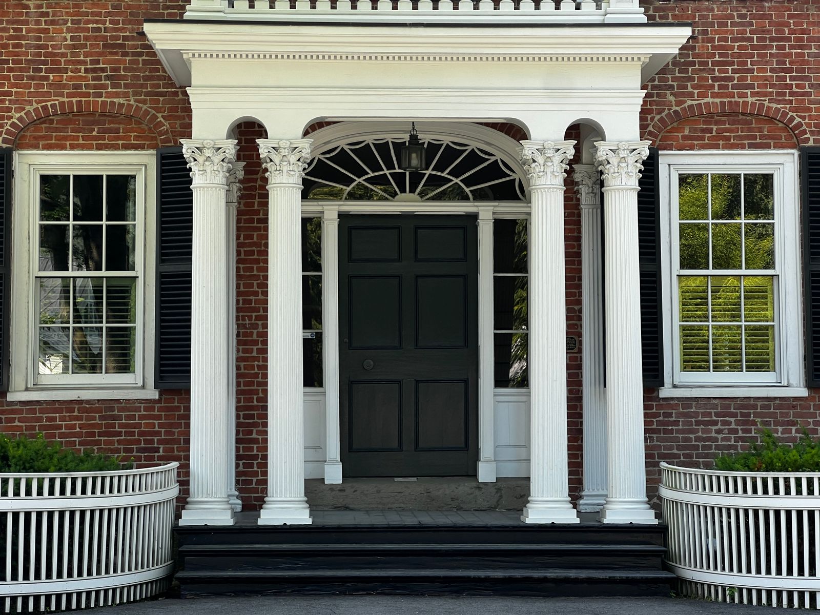 A columned white portico on an older brick house, framing a black paneled front door beneath a fanlight, with shuttered multi-pane windows on either side