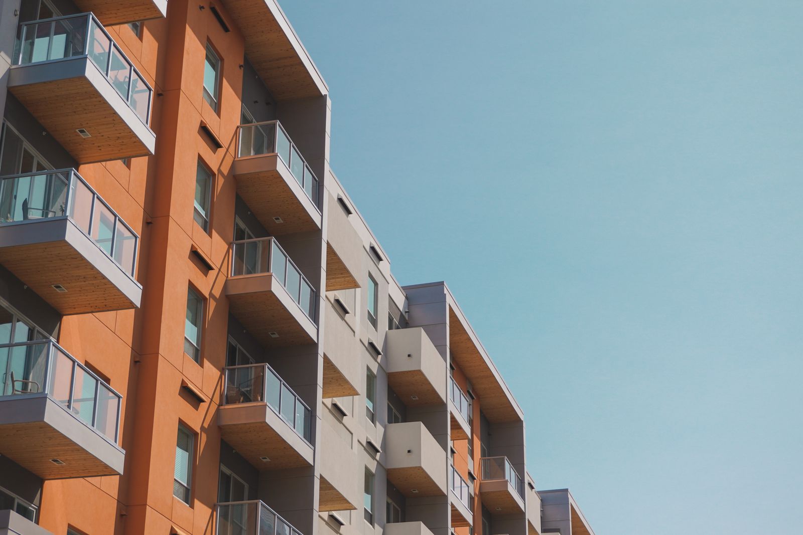 Upward view of a modern mid-rise condominium building with orange and gray stucco panels and glass-railed balconies stacked floor over floor against a clear blue sky