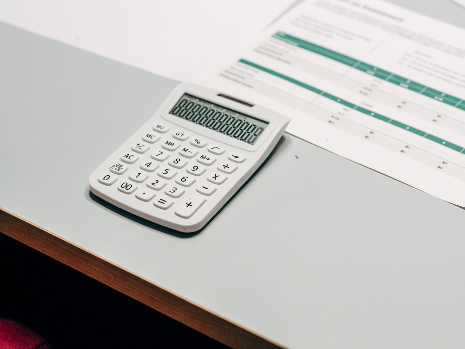 A white desktop calculator sitting on a pale desk beside a printed financial worksheet with green-shaded rows of figures