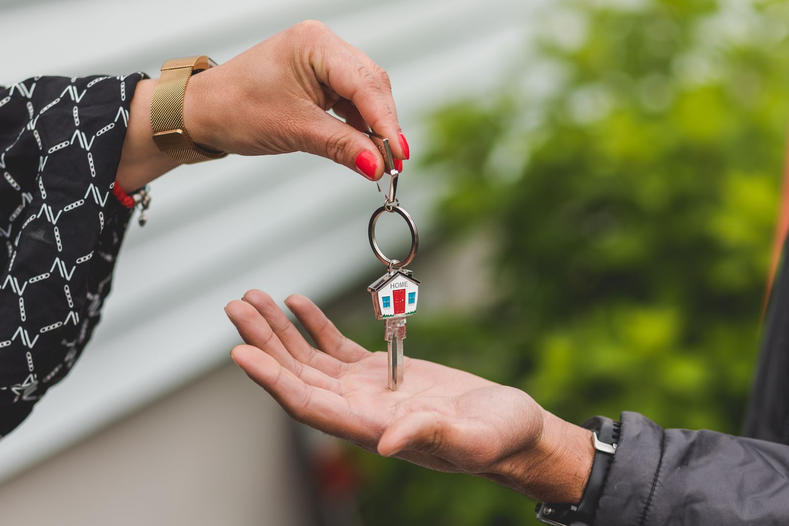 One hand dropping a house key on a home-shaped keychain into another person's open palm, outside a house with gray siding
