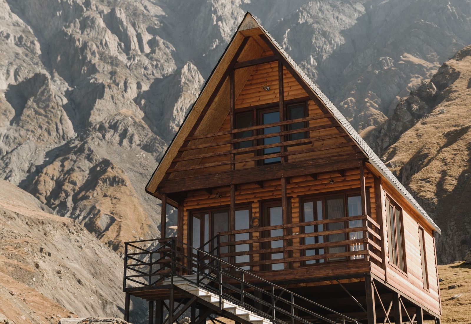 A two-story A-frame wooden cabin on stilts with a wraparound balcony and an exterior staircase, set on a dry rocky slope beneath steep gray mountains