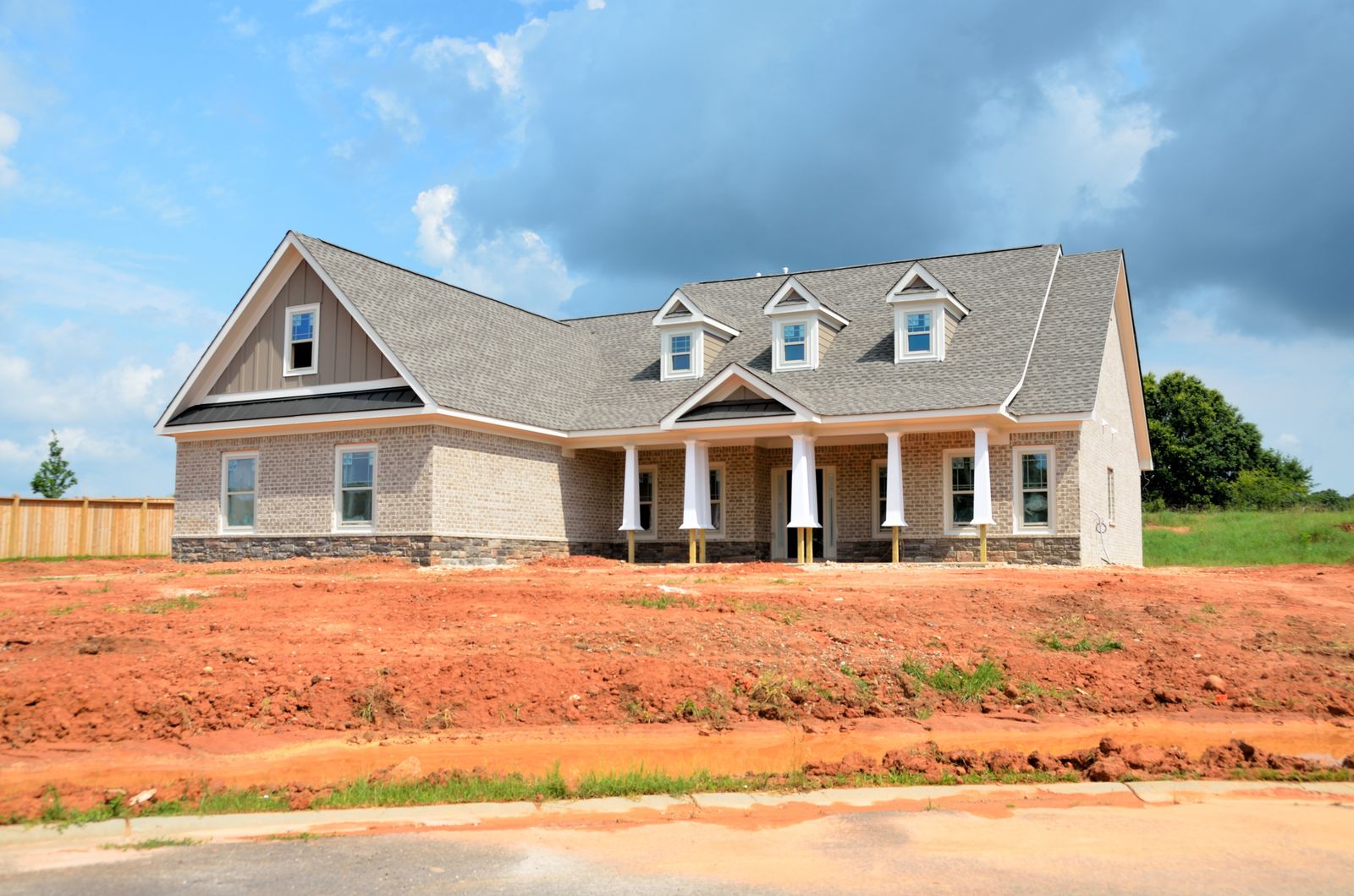A newly finished brick house with dormer windows and a columned front porch, standing on a bare graded dirt lot with no landscaping and a fresh sidewalk along the front