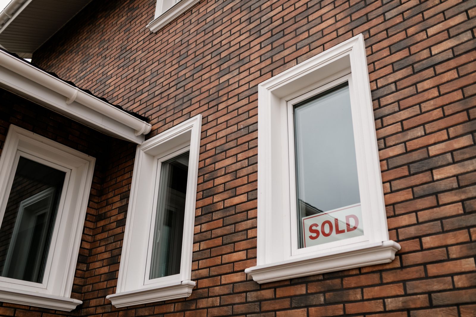 Close-up of a red brick two-story house with white-trimmed windows, a small red SOLD card taped inside one of the lower window panes